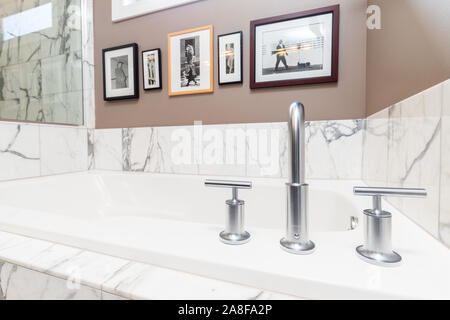 Detail shot of a fancy bathtub surrounded by white and gray marble with a chrome Kohler faucet and pictures hung on the wall. Stock Photo