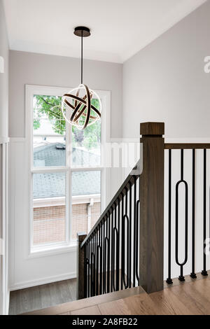 A stairway leading downstairs with wrought iron poles on the railings and a modern, curved light hanging from the ceiling with a colorful view outside Stock Photo