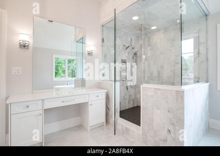 A bright luxury bathroom with a large shower and small, white vanity counter top. A stand alone bath tub can be seen through the mirror. Stock Photo