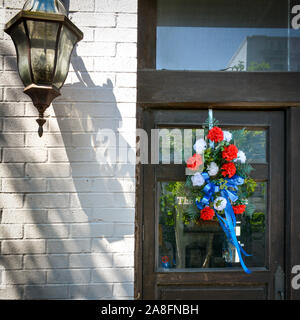 A red, white and blue memorial wreath hangs on an old wooden door with  window glass on  a historic building with a lantern along side on brick wall Stock Photo