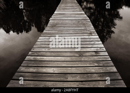 A mystical seeming wooden footbridge crosses the water at twilight with trees reflecting in water for a zen experience Stock Photo