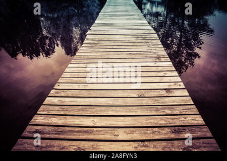 A mystical seeming wooden footbridge crosses the water at twilight with trees reflecting in water for a zen experience Stock Photo