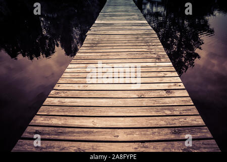 A mystical seeming wooden footbridge crosses the water at twilight with trees reflecting in water for a zen experience Stock Photo