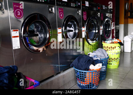 ANTIPOLO CITY, PHILIPPINES – NOVEMBER 2, 2019: Dirty clothes in a clothes basket in front of rows of washing machines inside a laundromat. Stock Photo