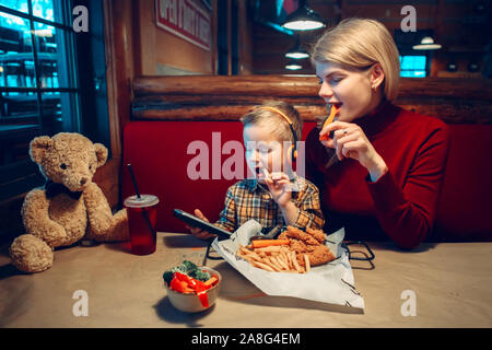 Caucasian family mother with toddler boy son eating food dinner in cafe restaurant and playing electronic digital gadget device toy. Children kids adu Stock Photo
