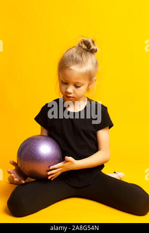 Young beautiful white caucasian girl gymnast doing gymnastic exercise with a sports ball on yellow background Stock Photo