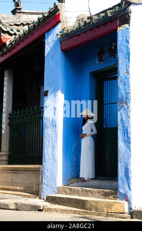 Young Vietnamese woman wearing a white ao dai  or traditional tunic with pants and a conical straw hat or non la. She is standing at the entrance to T Stock Photo