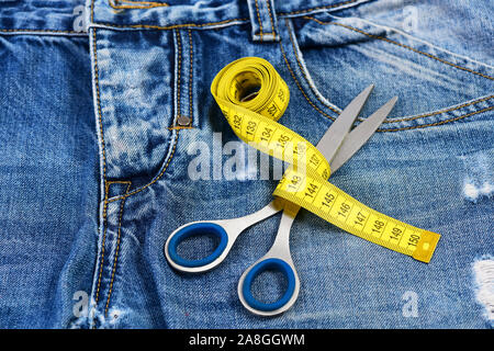 Tailors tools on denim fabric, selective focus. Measure tape wound around metal scissors on jeans. Jeans crotch, pocket and belt loops, close up. Maki Stock Photo