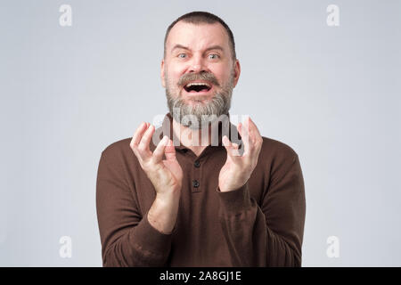 Mature scared bearded man in brown clothes squeezes fingers from horror. Negative facial emotion. Stock Photo
