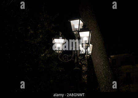 Four lighted electric lamps on a single lamp post at night. A moody dark shot with trees in the background Stock Photo
