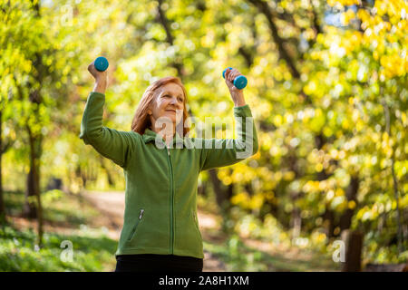 Senior woman is exercising with weights in park on sunny day. Stock Photo