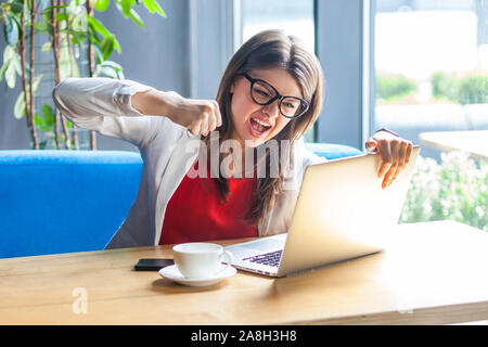 Portrait of angry stylish brunette young woman in glasses sitting and boxing, punching her laptop with crazy emotion. business problem concept. indoor Stock Photo
