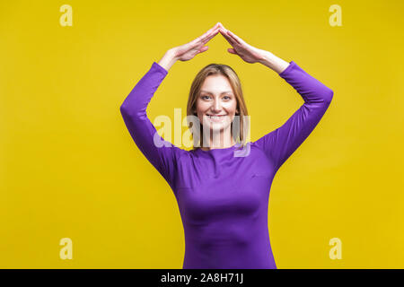 Secured life, insurance concept. Portrait of cheerful woman in elegant tight purple dress showing home roof gesture with hands over head, feels safe. Stock Photo