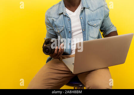 Cropped portrait of male photographer in denim shirt holding digital dslr camera and laptop, going to take picture or create video, using photo editin Stock Photo