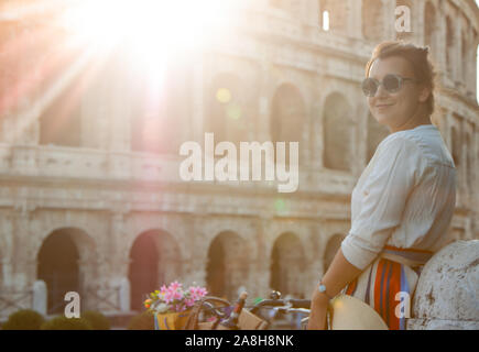 Beautiful young woman with bike in fashion dress standing in front of colosseum in Rome at sunset. Stock Photo