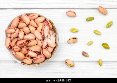 Overhead shot, roasted pistachios nuts in small bowl, some scattered on white boards desk. Stock Photo