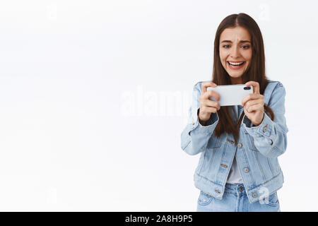 Distressed, intense young attractive woman in denim, holding smartphone horizontally, frowning and grimacing as losing online mobile game battle Stock Photo