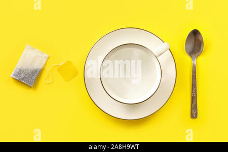 White porcelain cup filled with hot water, tea bag and spoon next to it on yellow board, photo from above. Stock Photo