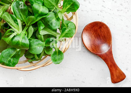 Small basket with corn salad (Valerianella locusta), tiny wooden spoon next to it on white board, photo from above. Healthy green leaves concept. Stock Photo