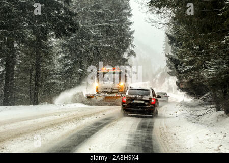 Driving on forest road through snowstorm, orange maintenance plough truck coming opposite way, view from car behind. Stock Photo