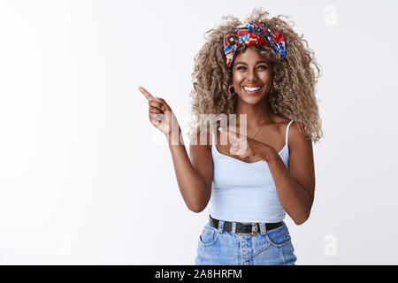 Cute sincere, friendly-looking blond curly-haired hipster woman with piercing, squinting joyfully, sharing cool link, pointing fingers left blank copy Stock Photo