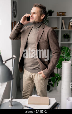 Confident mature architect with smartphone standing by office window Stock Photo