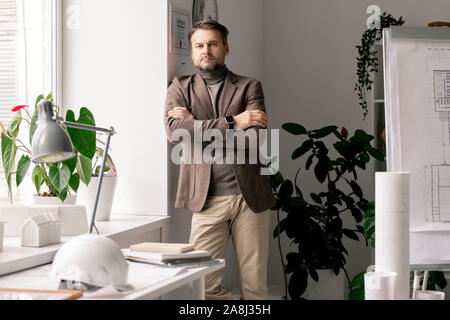 Serious architect or engineer in formalwear standing by workplace by window Stock Photo
