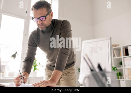 Confident engineer bending over papers while drawing new sketches in office Stock Photo