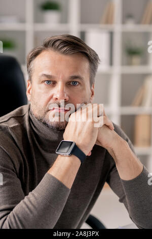 Serious mature man with smartwatch on right wrist sitting by workplace in office Stock Photo