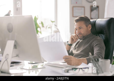 Mature serious architect looking at sketches while checking them by workplace Stock Photo