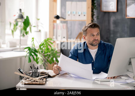 Serious engineer with blueprint looking through data on computer screen Stock Photo