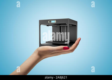 Side closeup of woman's hand facing up and holding black 3D printer on light blue background. Stock Photo