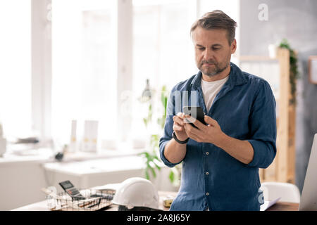 Serious engineer in casualwear scrolling through contacts in his smartphone Stock Photo