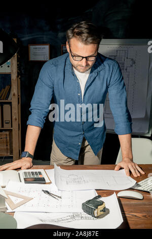 Mature engineer in casualwear looking at sketch on paper while working overtime Stock Photo