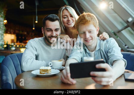 Two happy guys and pretty blonde girl making selfie on smartphone in cafe Stock Photo
