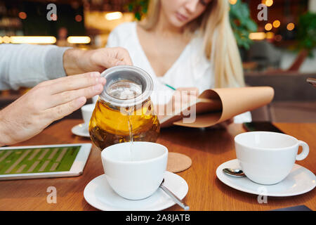 Hands of young man with teapot pouring fresh herbal tea into white porcelain cup Stock Photo