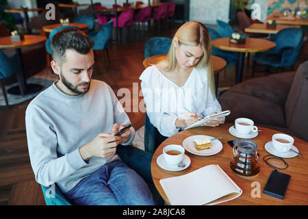Casual guy scrolling in gadget while his girlfriend making notes or sketch Stock Photo