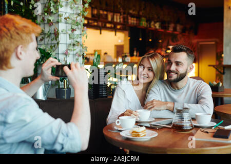 Young cheerful couple posing for their college friend with smartphone in cafe Stock Photo
