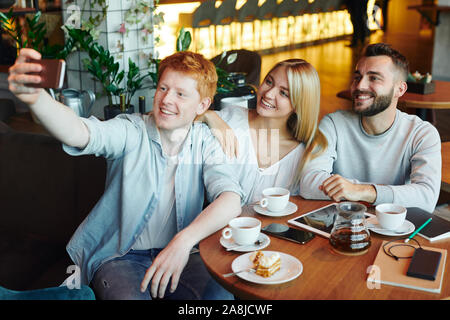 Three happy young affectionate friends making selfie in cozy cafe by cup of tea Stock Photo