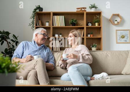 Young female and her father having tea while relaxing and chatting on couch Stock Photo