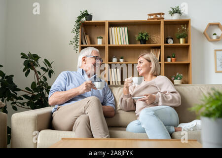 Happy female and her senior father having tea while sitting on couch and talking Stock Photo