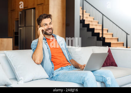 Leisure at Home. Young man sitting on sofa with laptop talking on phone smiling relaxed Stock Photo