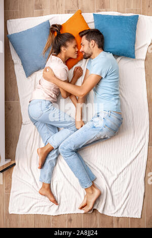Mixed Race Marriage. Young man hugging looking at sleeping woman happy lying on bed top view Stock Photo