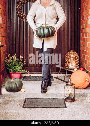 Unidentified woman standing on doorway, wearing cozy white knitted sweater and holding green muscat pumpkin in hands. Outdoor seasonal exterior home f Stock Photo