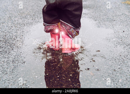 Girl having fun, jumping in water puddle on wet street, wearing rain boots with reflective detail fabric stripes shining. High visibility and safety i