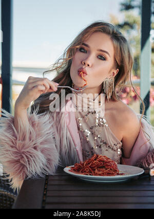 Cute girl sitting in a restaurant and eating pasta (spaghetti). Portrait of stylish young woman in cafe, wearing dress and fur coat. Film grain effect Stock Photo