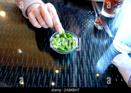 A glass bowl with shelled edamame soy beans on a restaurant table top with a woman's hand reaching in to take one. Stock Photo