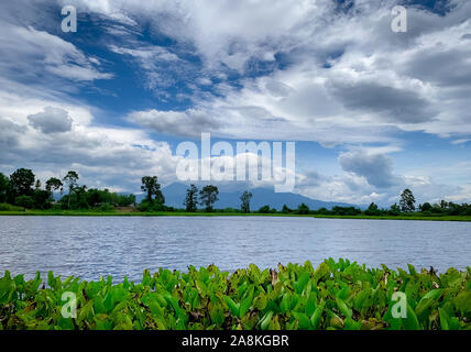 Beautiful landscape view of lake in front of the mountain with blue sky and white cumulus clouds. Green tree and grass field around pond. Tropical Stock Photo
