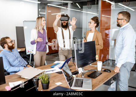 Group of diverse colleagues during small conference in the office, creative bearded man trying a new product with virtual reality goggles Stock Photo