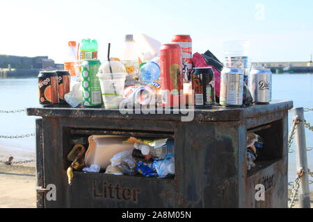 Overflowing rubbish bin / trash can / litter bin at the seaside with empty cans, plastic bottles and other trash on top. Illustrating not recycling. Stock Photo